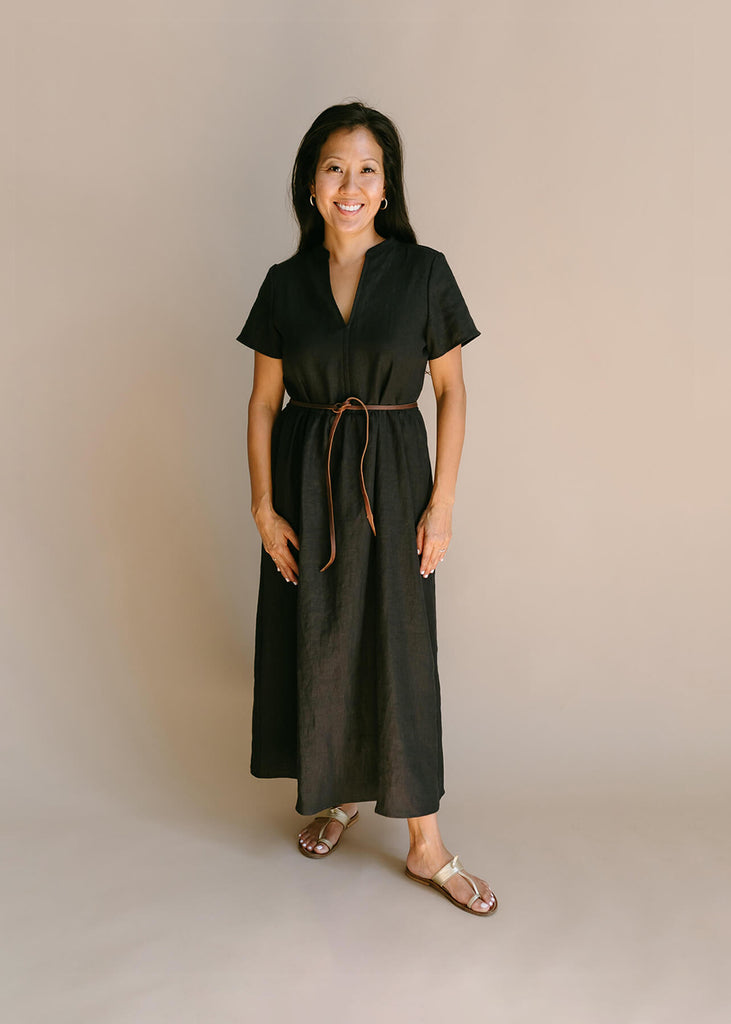 a woman wearing a black, 100% linen, ankle length, v-neckline dress with fabric gathered at the waist, and two pockets and a belt.