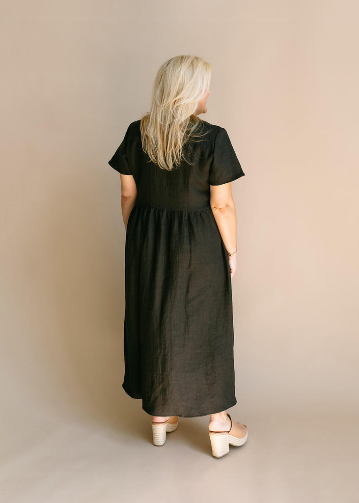 Back view of a woman wearing a black, 100% linen, ankle length, v-neckline dress with fabric gathered at the waist, and two pockets.
