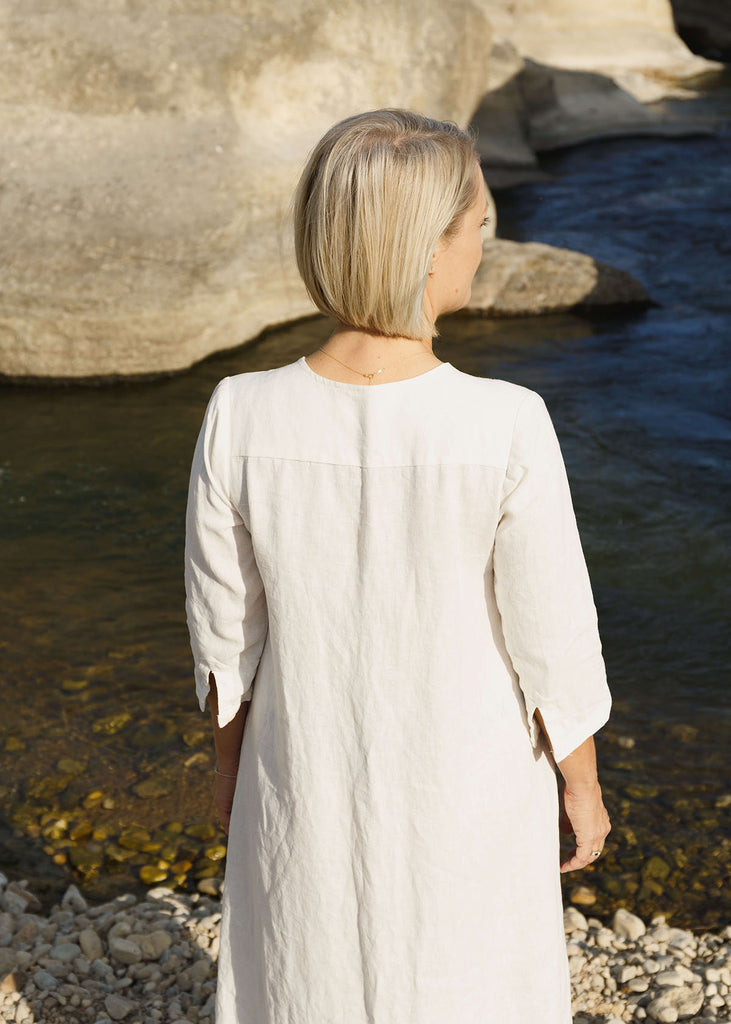Back view of a woman wearing the Sienna everyday buttoned shirtdress in white, with ¾ sleeves, a V-neck, an uneven hemline.