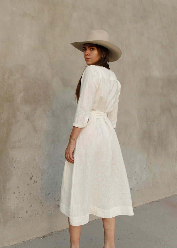 Back view of a woman wearing the Sienna everyday buttoned shirtdress in white, with ¾ sleeves, a V-neck, an uneven hemline, with a belt.