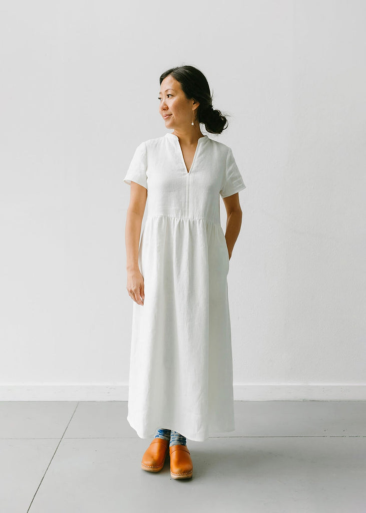 a woman wearing a white, 100% linen, ankle length, v-neckline dress with fabric gathered at the waist, and two pockets.
