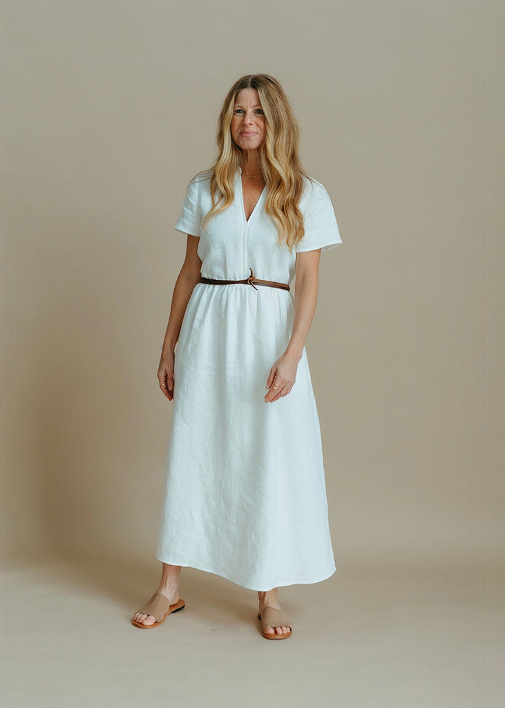 a woman wearing a white, 100% linen, ankle length, v-neckline dress with fabric gathered at the waist, and two pockets and a belt.