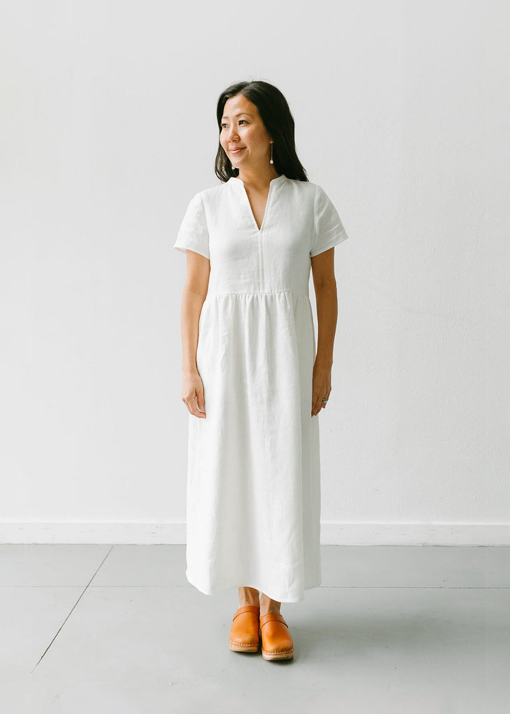 a woman wearing a white, 100% linen, ankle length, v-neckline dress with fabric gathered at the waist, and two pockets.