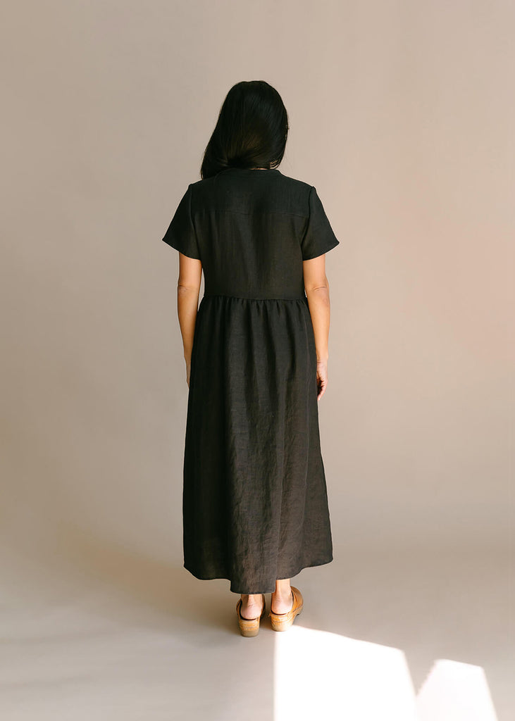 Back view of a a woman wearing a black, 100% linen, ankle length, v-neckline dress with fabric gathered at the waist, and two pockets.