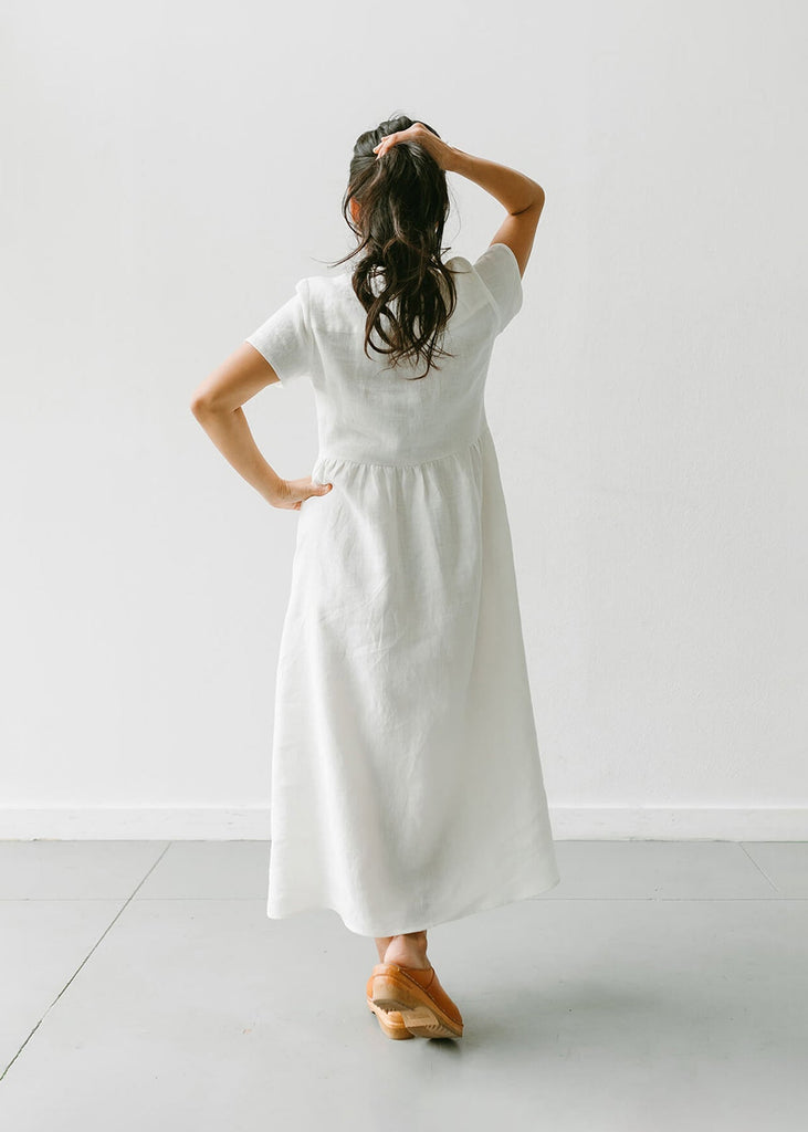 back view of a woman wearing a white, 100% linen, ankle length, v-neckline dress with fabric gathered at the waist, and two pockets.