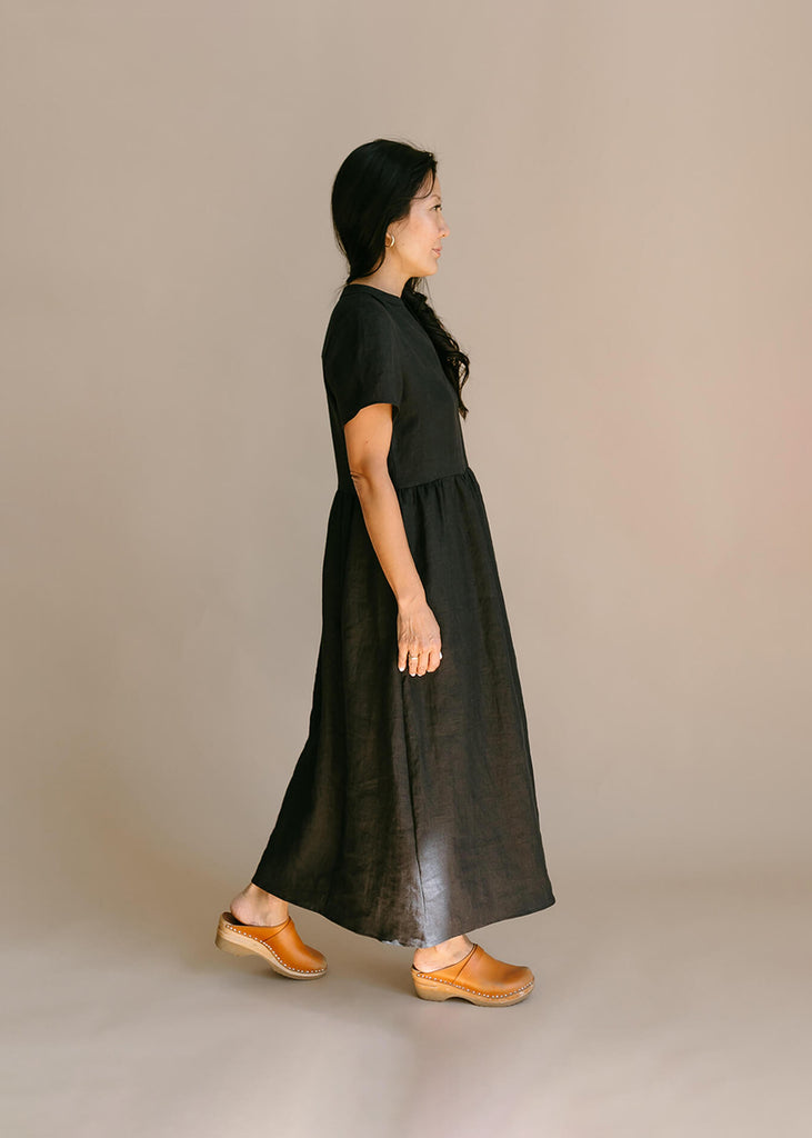 Side view of a woman wearing a black, 100% linen, ankle length, v-neckline dress with fabric gathered at the waist, and two pockets.