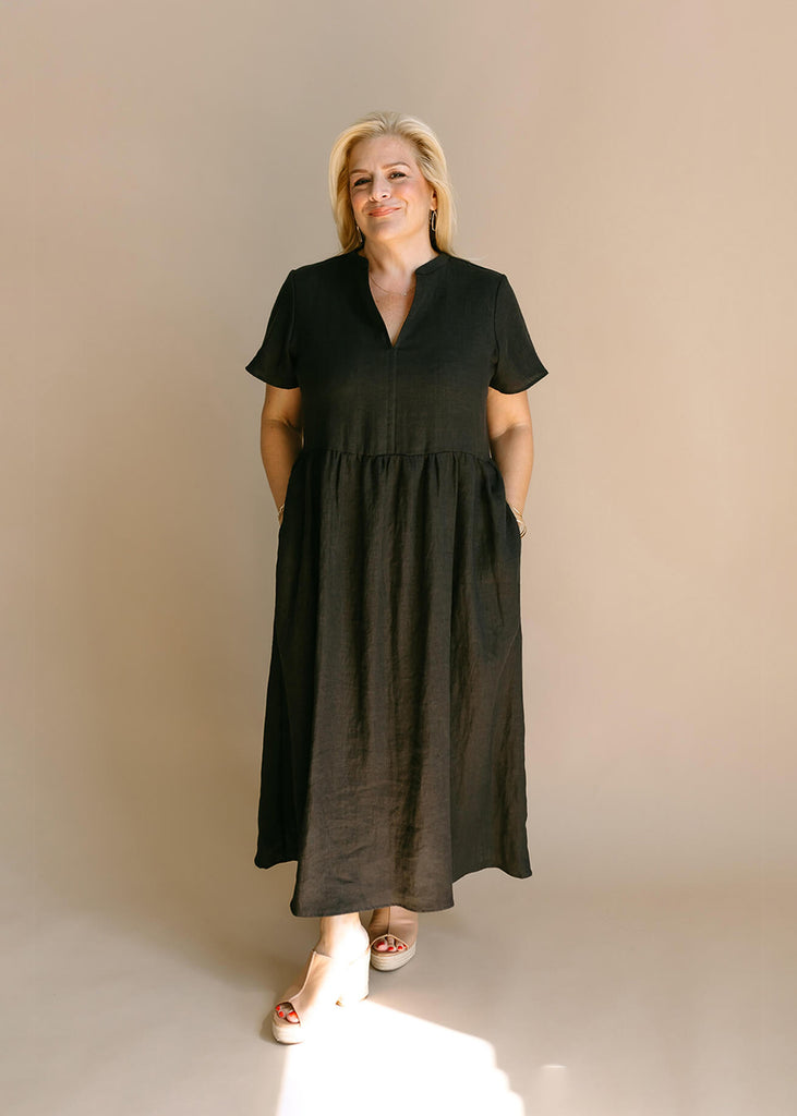 a woman wearing a black, 100% linen, ankle length, v-neckline dress with fabric gathered at the waist, and two pockets.