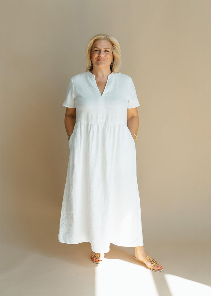 a woman wearing a white, 100% linen, ankle length, v-neckline dress with fabric gathered at the waist, and two pockets.