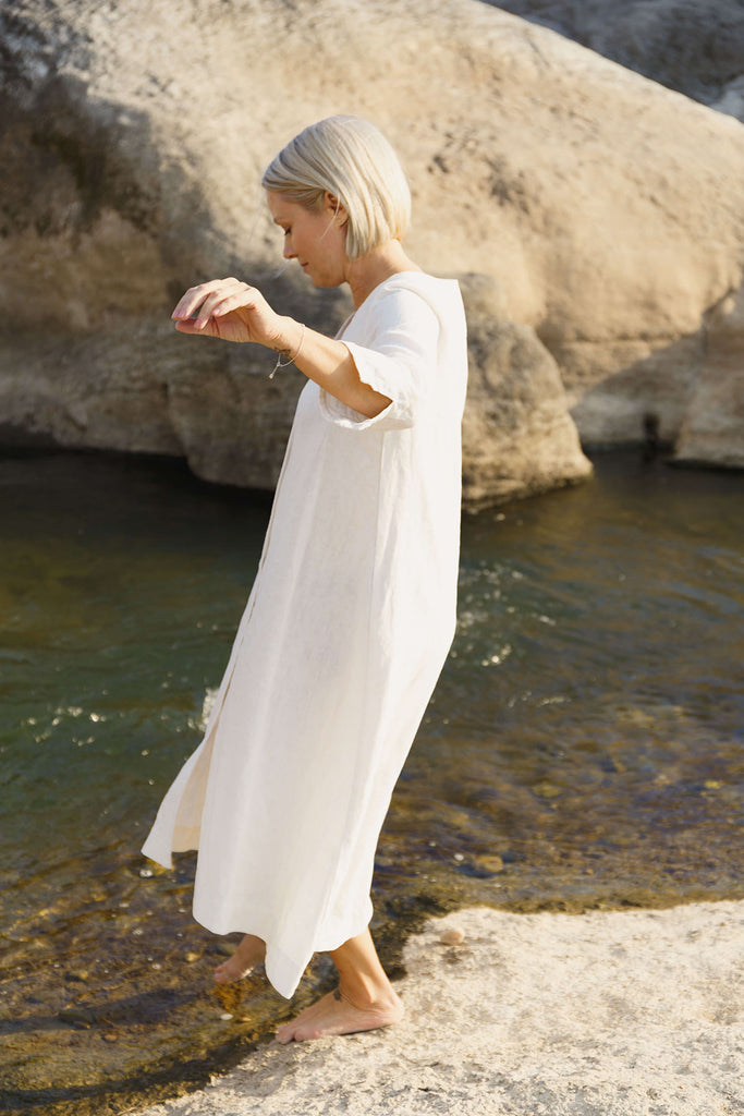woman stepping into a creek wearing The Sienna Dress: everyday shirtdress with ¾ sleeves, a V-neck, an uneven hemline, made of 100% ramie.