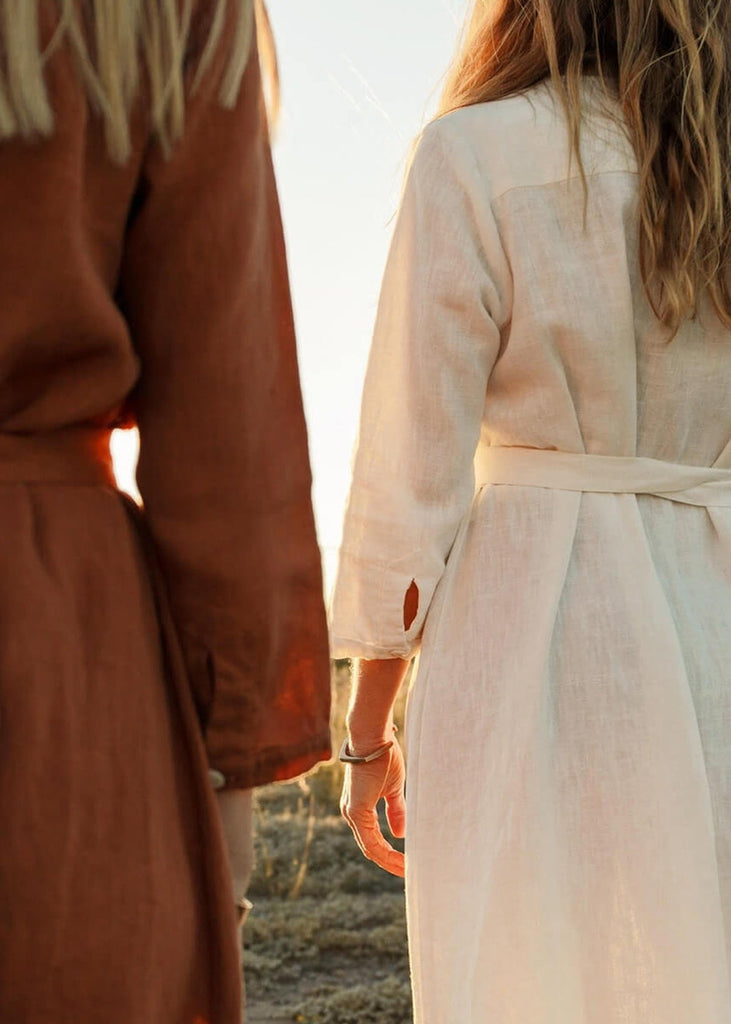 Back view of two women wearing the Sienna everyday buttoned shirtdress in burnt sienna and one in white, with ¾ sleeves, a V-neck, an uneven hemline, with a belt.