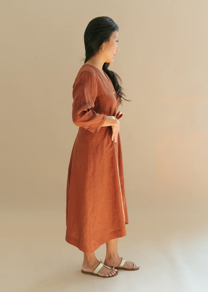 Side view of a woman wearing the Sienna everyday buttoned shirtdress in burnt sienna with ¾ sleeves, a V-neck, an uneven hemline, with a belt.
