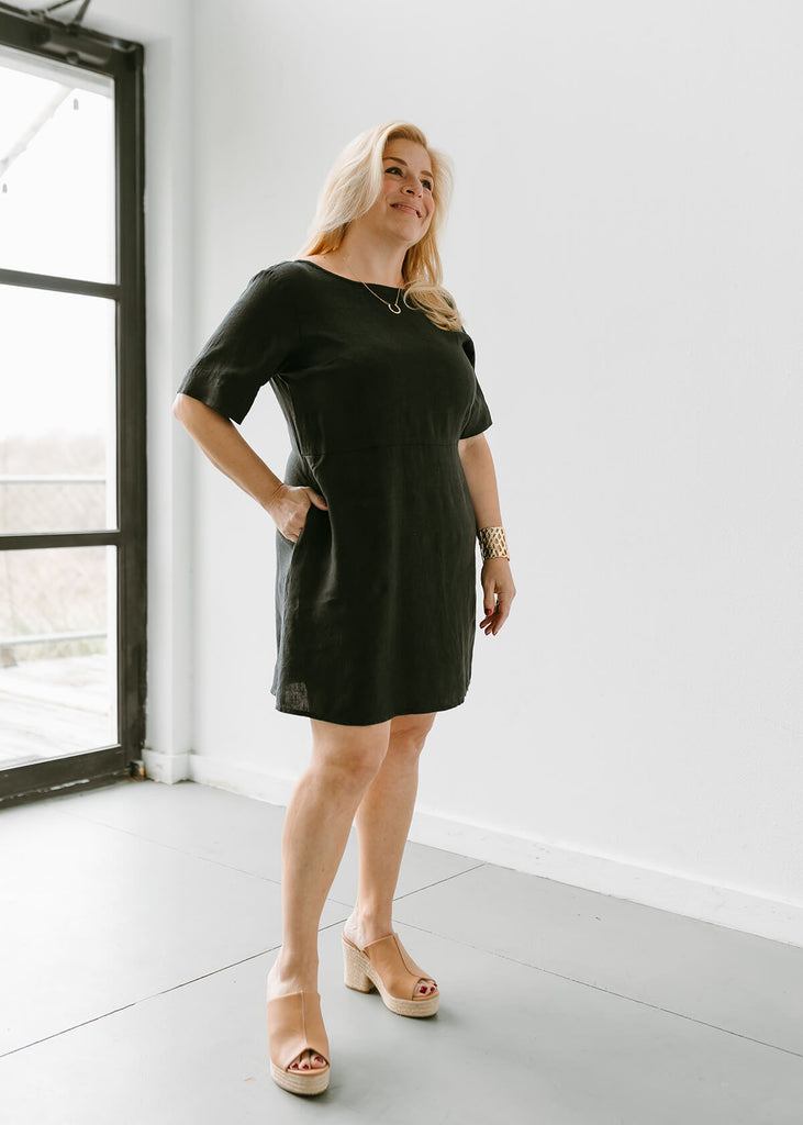 side view of woman wearing a linen dress that hits above the knee, all black with three quarter length sleeves and two pockets.