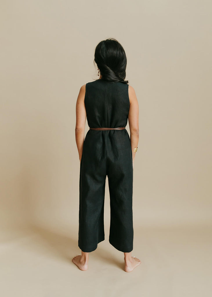 back of woman wearing a linen jumpsuit with a loose bodice with a V neck front zip opening, a detached self tie belt, one front pocket and wide cropped leg.