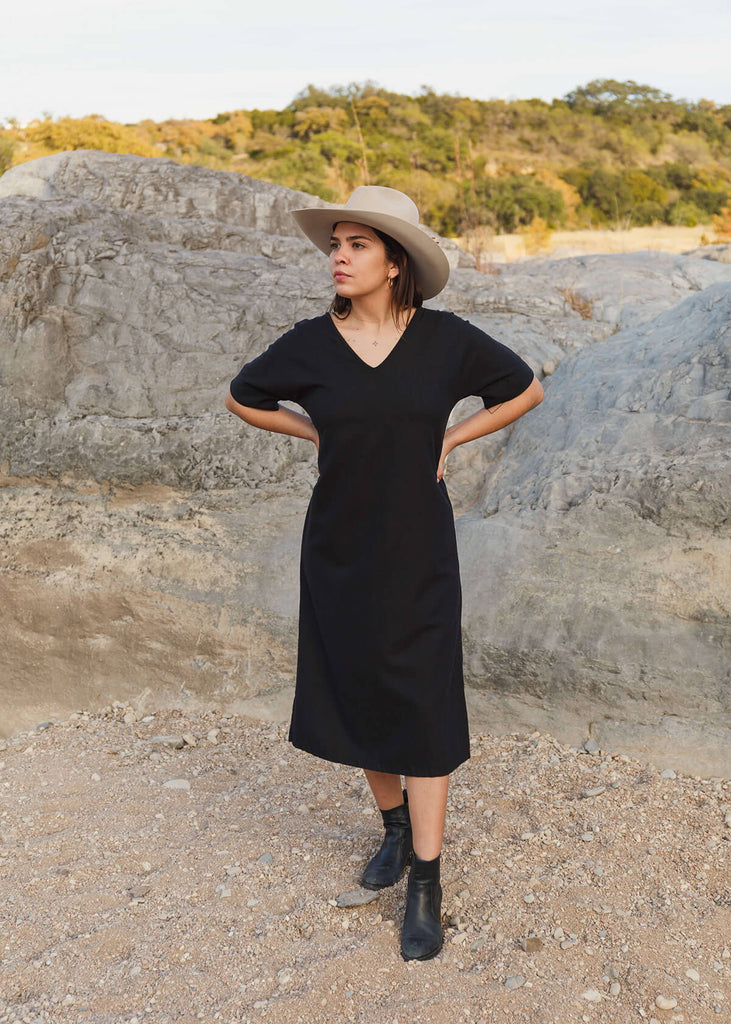 woman standing with hands on her hips wearing a mid-calf length black dress with three-quarter length sleeves. 