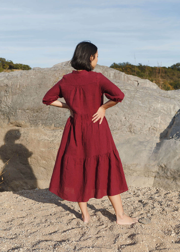 Back view of a woman wearing a 100% Japanese linen tiered dress with a loose fit and gathering at the waist, in a burgundy color.