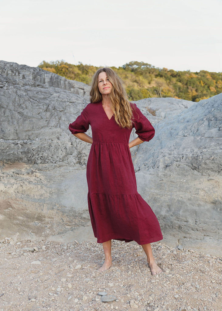 A woman wearing a 100% Japanese linen tiered dress with a loose fit and gathering at the waist, in a burgundy color.