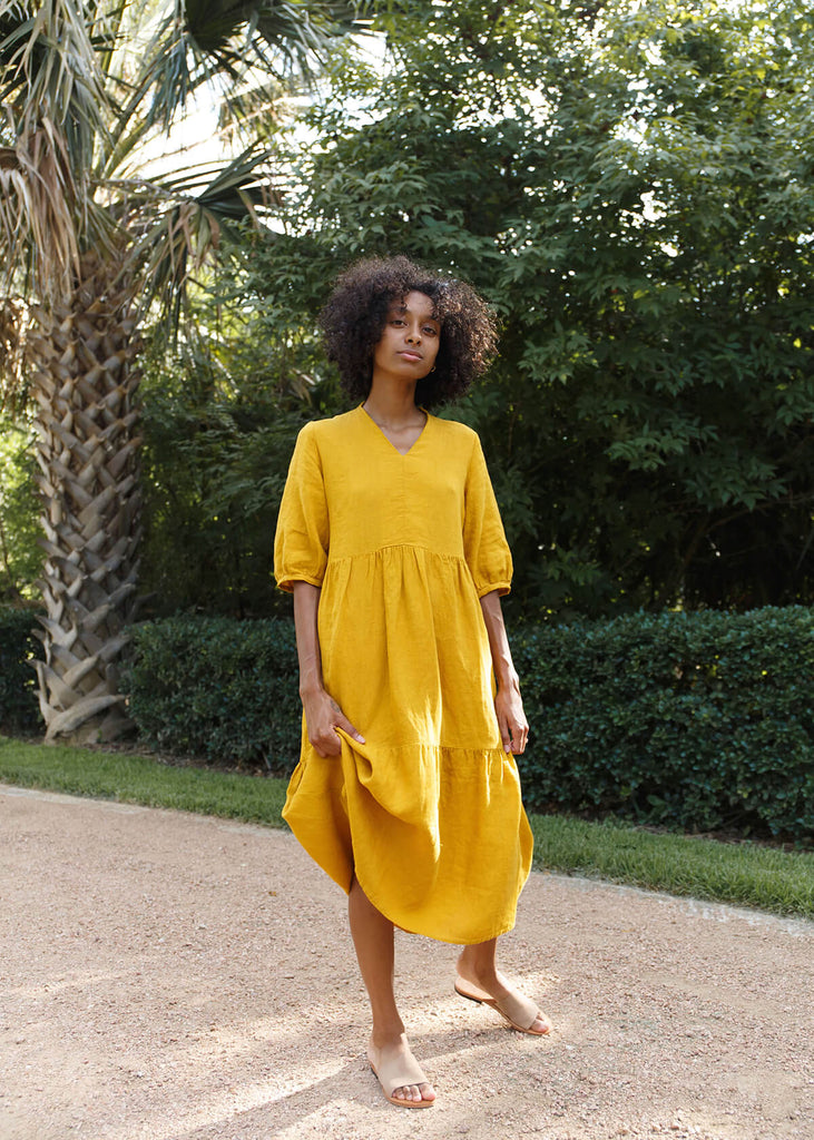  A woman wearing a 100% Japanese linen tiered dress with a loose fit and gathering at the waist, in a bright yellow color.
