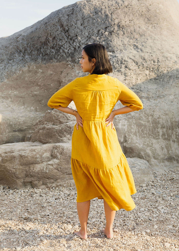  Back view of a woman wearing a 100% Japanese linen tiered dress with a loose fit and gathering at the waist, in a yellow color.