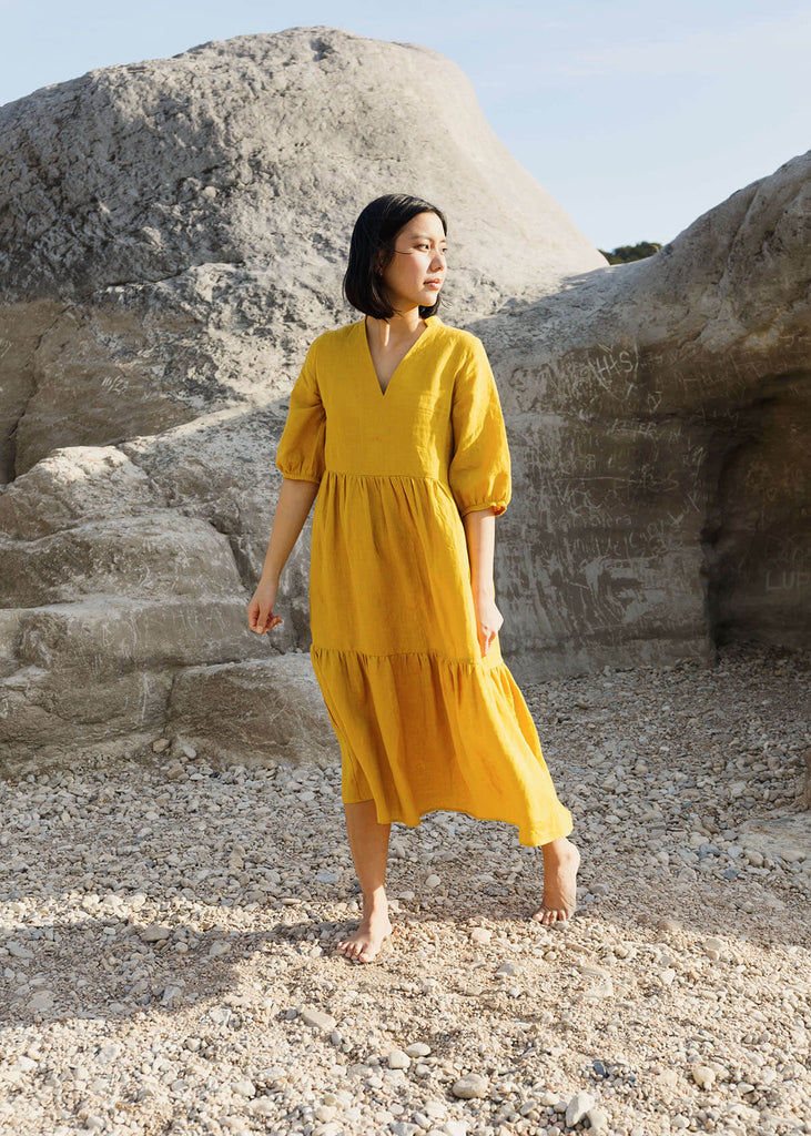  A woman wearing a 100% Japanese linen tiered dress with a loose fit and gathering at the waist, in a bright yellow color.