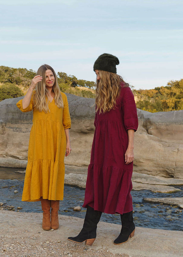 Two women, one pictured from the side, wearing a 100% Japanese linen tiered dress with a loose fit and gathering at the waist, in a bright yellow and burgundy color.
