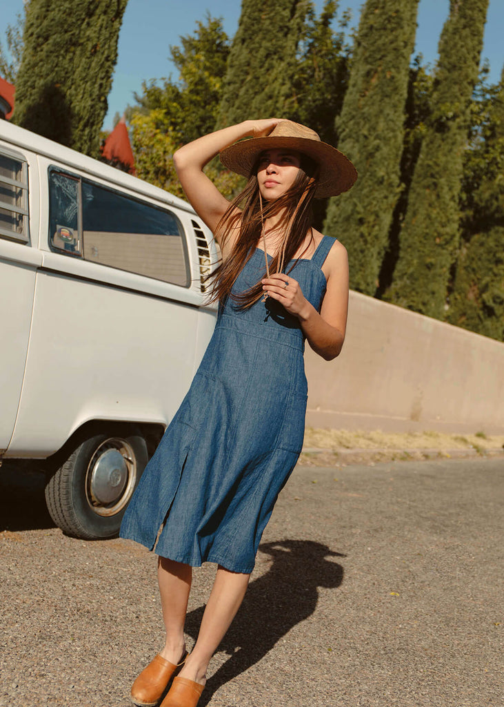 woman wearing a blue sundress with a fitted bodice and flattering A-line skirt; adjustable button straps, two pockets, one side slit.