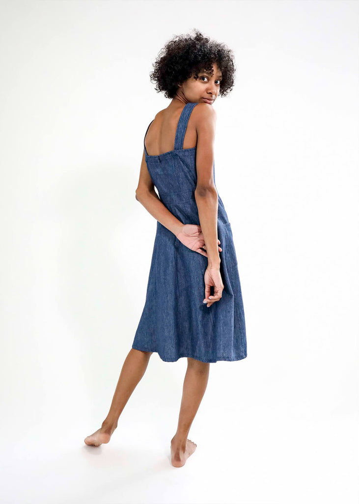 back view of a woman wearing a blue sundress with a fitted bodice and flattering A-line skirt; adjustable button straps, two pockets, one side slit.