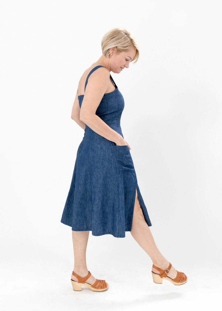 side view of a woman wearing a blue sundress with a fitted bodice and flattering A-line skirt; adjustable button straps, two pockets, one side slit.