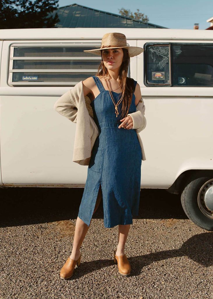 woman wearing a blue sundress with a fitted bodice and flattering A-line skirt; adjustable button straps, two pockets, one side slit.