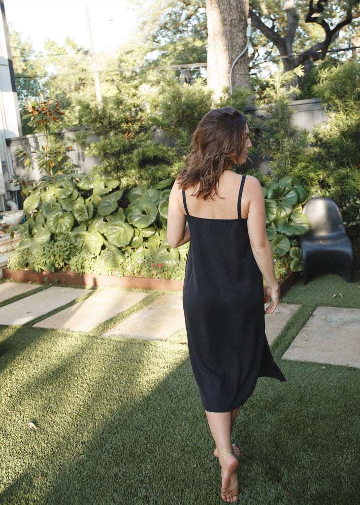 back view of  a woman wearing a black, 100% Tencel relaxed, straight fit slip dress with two side slits