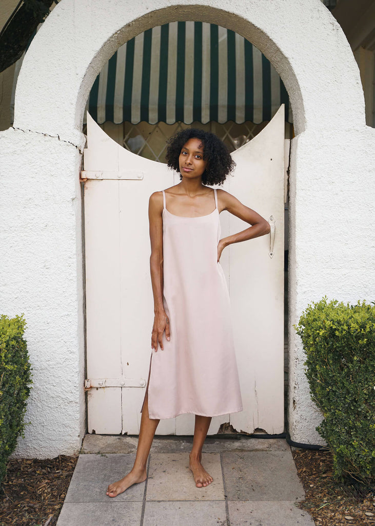  a woman wearing a blush-colored, 100% Tencel relaxed, straight fit slip dress with two side slits