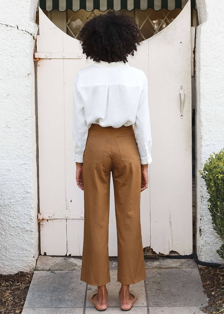 back view of woman wearing brown high-waisted, wide-leg pants with a slight ankle crop, single button front closure, and back pockets.