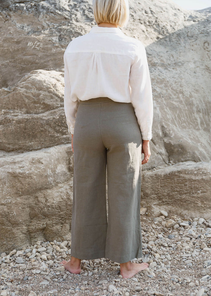 back view of a woman wearing natural gray colored pants in a wide-leg, slightly cropped style with no pockets, and Zipper and single button front closure.