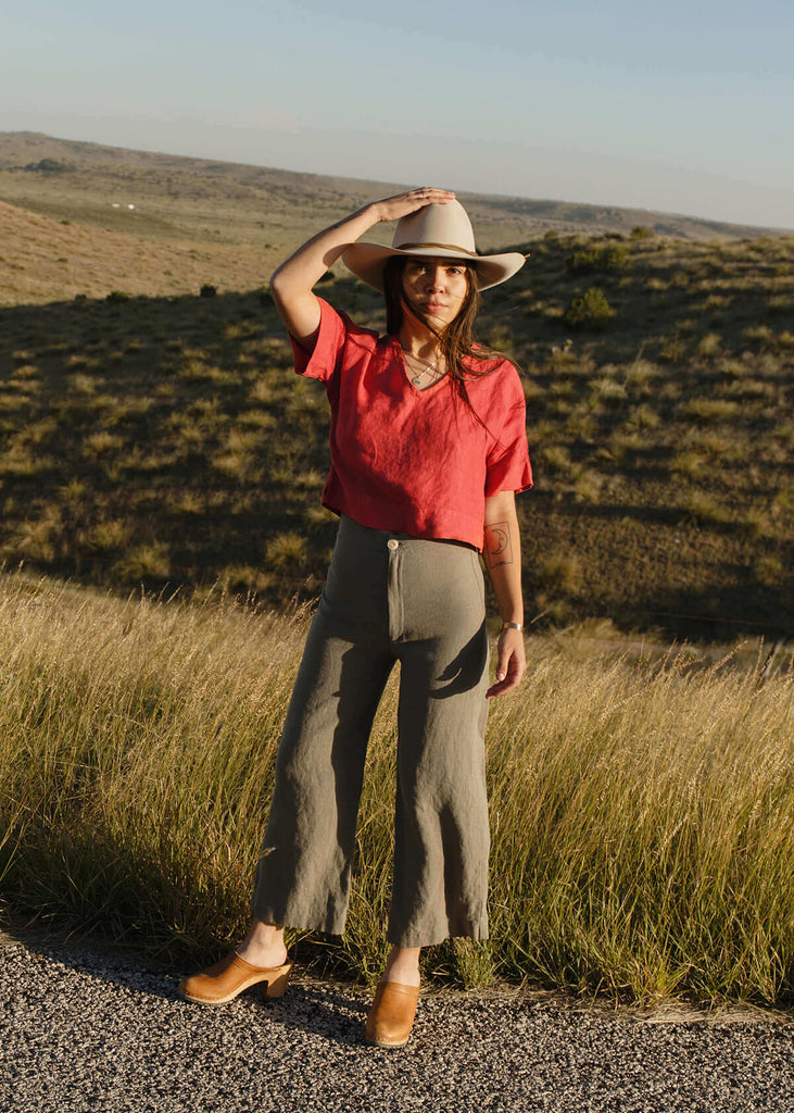 Woman wearing a boxy crop top with V neck front, relaxed dropped shoulder sleeves, two inch hems and two small side slits, in a coral red color.