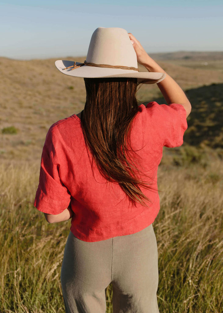 Back view of Woman wearing a boxy crop top with V neck front, relaxed dropped shoulder sleeves, two inch hems and two small side slits, in a coral red color.