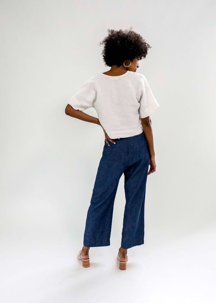 Back view of Woman wearing a boxy crop top with V neck front, relaxed dropped shoulder sleeves, two inch hems and two small side slits, in white.